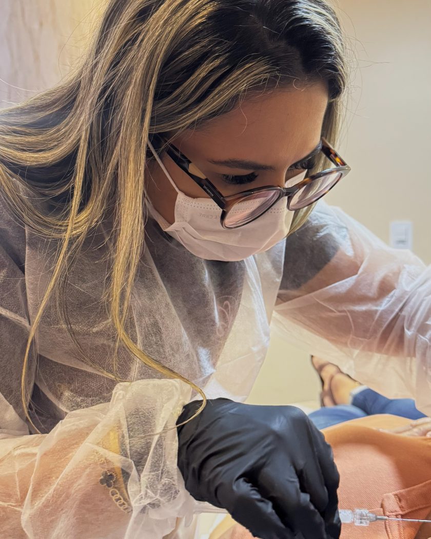 Dra. Najara Vieira de máscara, óculos e avental descartável aplicando bioestimulador de colágeno com cânula, com luvas pretas